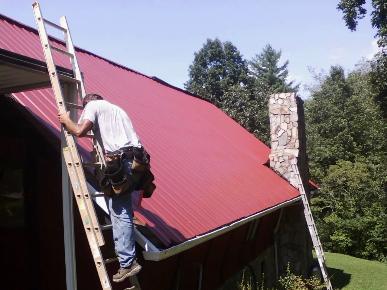 Roofer on a ladder beside a red metal roof and stone chimney — Soffit & Fascia Repair by Roofing Brothers in Washougal